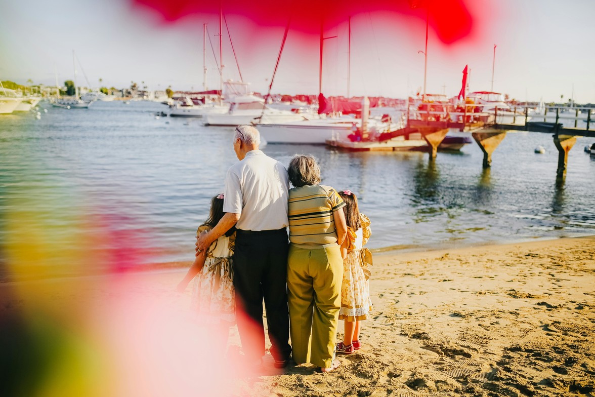Two grandparents and two young girls stand arm in arm on a sandy beach, looking out at a marina with sailboats in Ontario. Multigenerational family moment with warm sunlight and vibrant colour flares